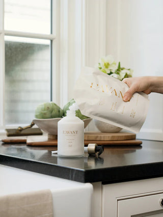 Person pouring a product from a white container labeled 'L'AVANT' into a white bottle on a kitchen counter. L'AVANT Fresh Linen Hand Lotion Refill Pouch