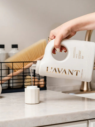 Person pouring laundry detergent from a L'AVANT container into a small container on a kitchen counter.