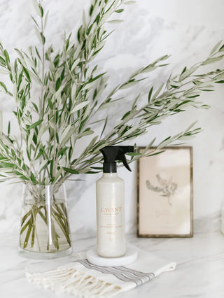 White bottle of Lavant fresh linen multipurpose spray with black sprayer on a white surface with greenery and a framed picture in the background.