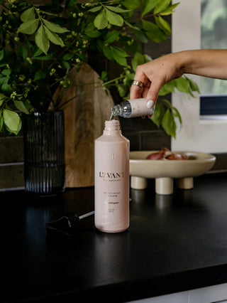 Person pouring a liquid into a pink bottle labeled 'L'evant' on a dark surface with greenery in the background. L'AVANT Blushed Bergamot Multipurpose Cleaner Refill