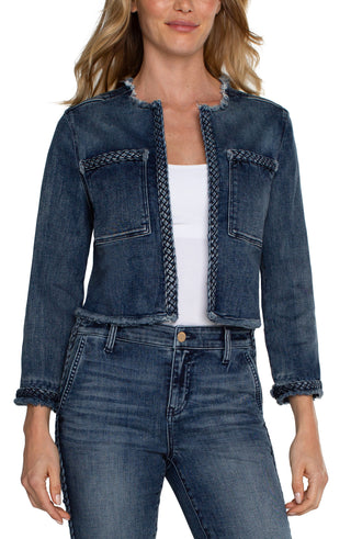 A woman wearing a cropped denim jacket with braided detailing, featuring a collarless design, frayed hem, and front patch pockets. She is also wearing a white top and blue jeans.