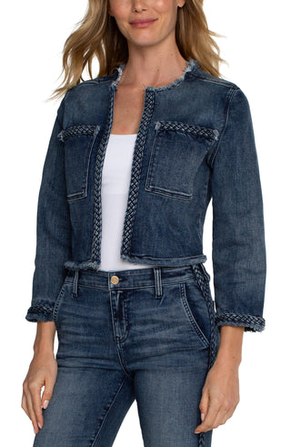 A woman wearing a cropped denim jacket with braided detailing, featuring a collarless design, frayed hem, and front patch pockets. She is also wearing a white top and blue jeans.