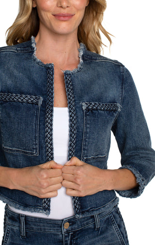 A woman wearing a cropped denim jacket with braided detailing, featuring a collarless design, frayed hem, and front patch pockets. She is also wearing a white top and blue jeans.