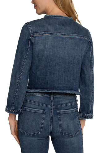 A woman wearing a cropped denim jacket with braided detailing, featuring a collarless design, frayed hem, and front patch pockets. She is also wearing a white top and blue jeans.