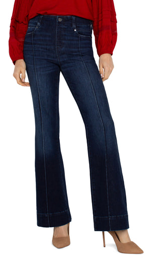 A pair of dark wash flare jeans with pintucks, featuring a mid-rise waist, 5-pocket styling, and a zip fly with a single button closure. The jeans are modeled with a heel.