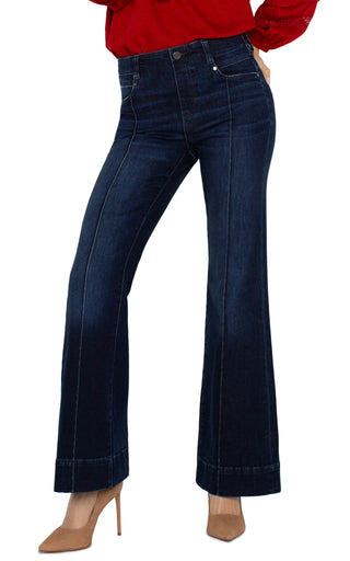 A pair of dark wash flare jeans with pintucks, featuring a mid-rise waist, 5-pocket styling, and a zip fly with a single button closure. The jeans are modeled with a heel.