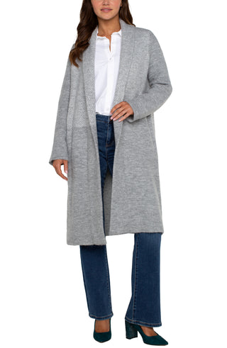 A woman wearing a long sleeve grey pattern open front shawl sweater coatigan with a white top and blue jeans.