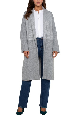 A woman wearing a long sleeve grey pattern open front shawl sweater coatigan with a white top and blue jeans.