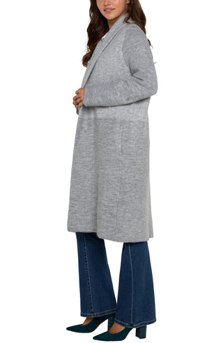 A woman wearing a long sleeve grey pattern open front shawl sweater coatigan with a white top and blue jeans.