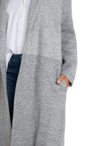 A woman wearing a long sleeve grey pattern open front shawl sweater coatigan with a white top and blue jeans.