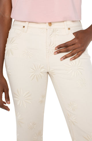 A pair of ecru colored crop flare jeans with floral embroidery and a frayed hem, featuring a mid-rise waist and a pair of tan sandals.