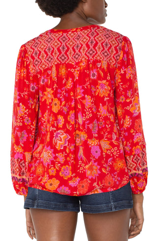 A woman wearing a red orange floral long sleeve button front shirred blouse with a V-neck.