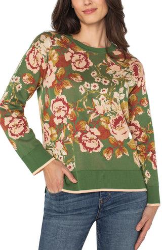 Woman wearing a green floral-patterned sweater with blue jeans on a white background