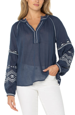 A woman wearing a long sleeve woven blouse in insignia blue with an embroidered pattern, paired with white pants.