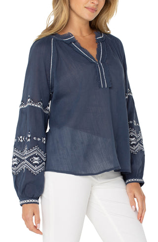 A woman wearing a long sleeve woven blouse in insignia blue with an embroidered pattern, paired with white pants.
