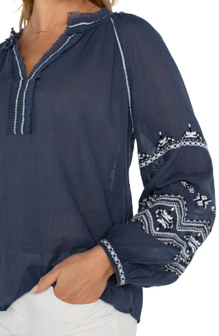 A woman wearing a long sleeve woven blouse in insignia blue with an embroidered pattern, paired with white pants.