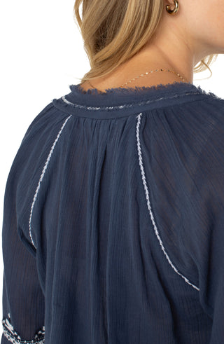 A woman wearing a long sleeve woven blouse in insignia blue with an embroidered pattern, paired with white pants.