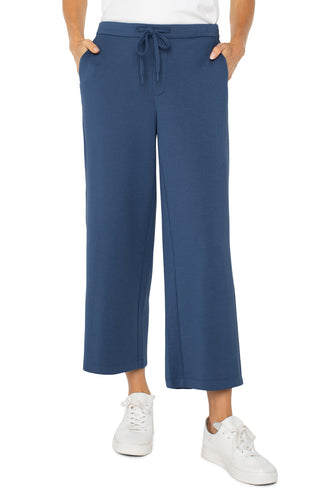 A person standing and wearing the Liverpool Pull On Wide Leg Crop Trouser in Prussian Blue with white sneakers.