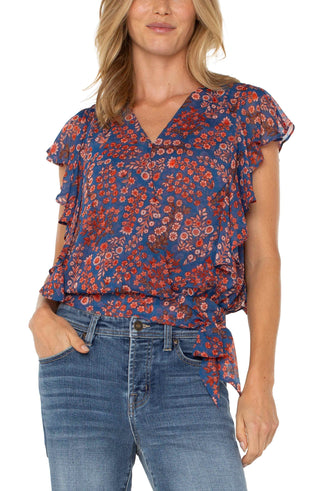 A woman wearing a Liverpool brand short sleeve blouse with a floral print, draped front, and waist tie. The blouse is paired with blue jeans.