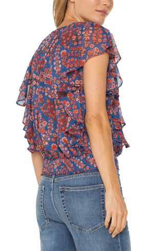 A woman wearing a Liverpool brand short sleeve blouse with a floral print, draped front, and waist tie. The blouse is paired with blue jeans.