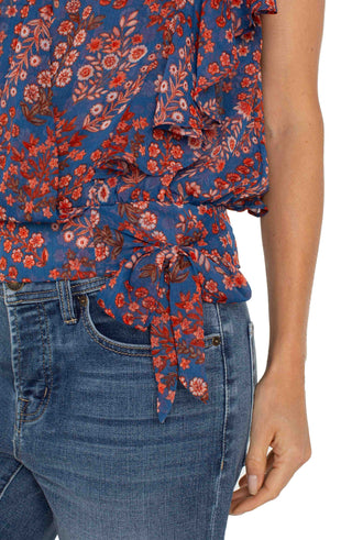 A woman wearing a Liverpool brand short sleeve blouse with a floral print, draped front, and waist tie. The blouse is paired with blue jeans.