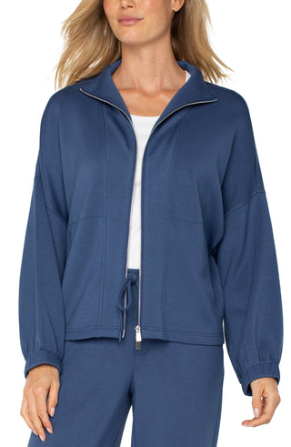 A woman wearing a blue zip up funnel neck jacket with a white top and blue trousers.