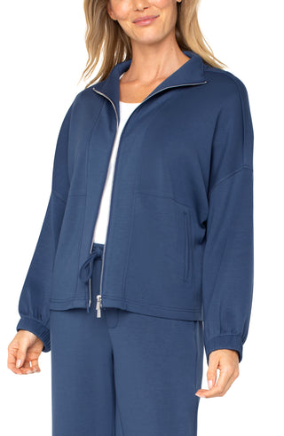 A woman wearing a blue zip up funnel neck jacket with a white top and blue trousers.