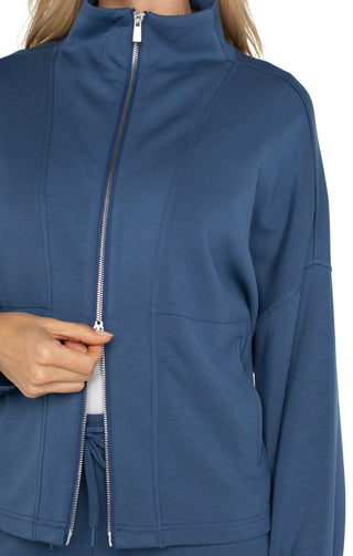 A woman wearing a blue zip up funnel neck jacket with a white top and blue trousers.