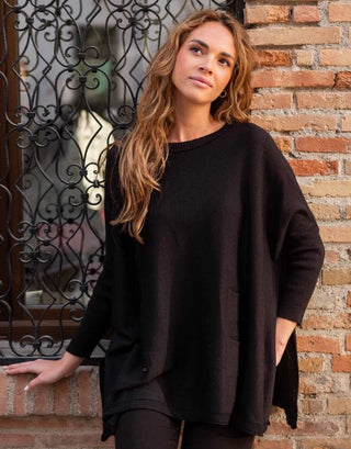 A woman standing by a window, wearing a black crewneck sweater with three pockets and split-sides design. Mersea Catalina Crewneck Sweater in Black