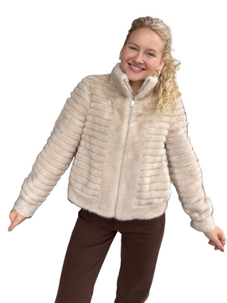 A woman modeling a cropped, ribbed faux fur jacket in a light beige color, paired with brown pants, against a plain white background. cream