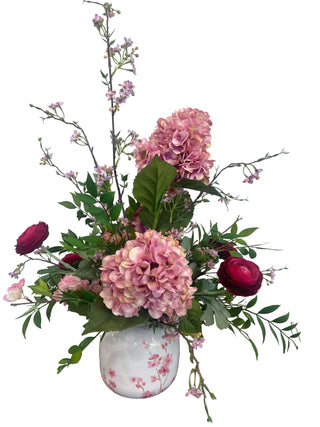 Pink Hydrangea & Cherry Blossom Arrangement 
A floral arrangement featuring pink hydrangeas, burgundy roses, and green foliage in a white ceramic vase with pink floral patterns