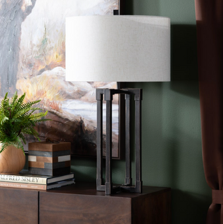 Table lamp with a white shade on a wooden surface next to books and a plant, with a painting in the background.