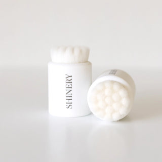 A white jewelry cleaning brush with a soft, tip and a white pill-shaped container with the brand name 'Shinery' printed on it.