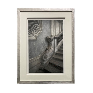A framed and matted artwork featuring a staircase with intricate details and a decorative railing, presented in a gray frame.