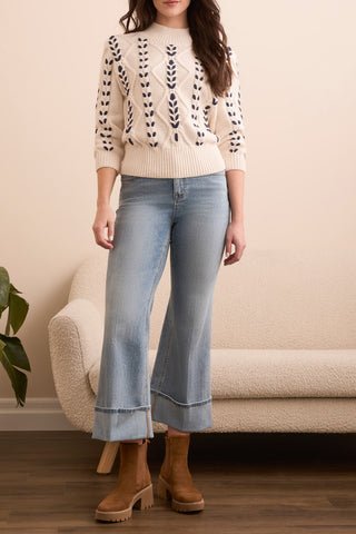 Woman wearing a cream sweater with navy weave patterns, light blue jeans, and brown boots in a living room setting.
