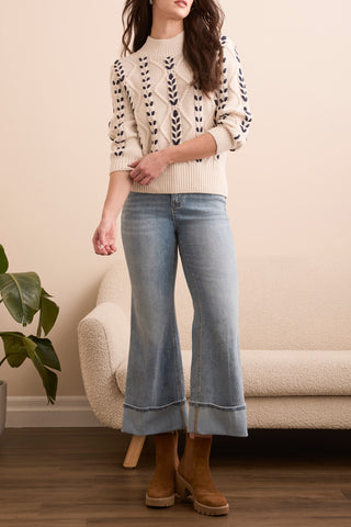 Woman wearing a cream sweater with navy weave patterns, light blue jeans, and brown boots in a living room setting.