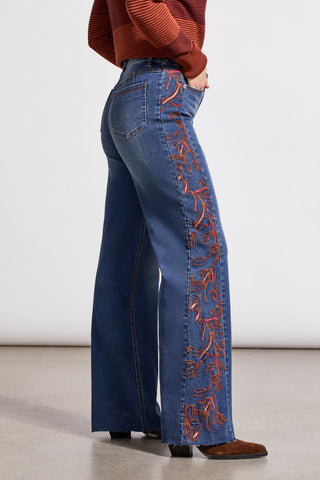 A person wearing blue denim jeans with intricate embroidery on the side, standing against a neutral background.