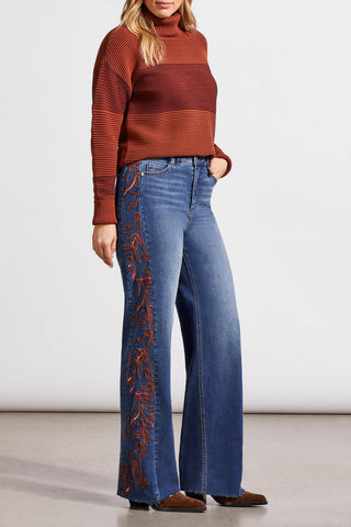 A person wearing blue denim jeans with intricate embroidery on the side, standing against a neutral background.