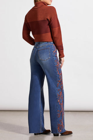 A person wearing blue denim jeans with intricate embroidery on the side, standing against a neutral background.