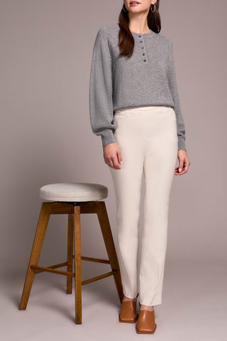 Woman wearing a gray sweater and white pants standing next to a wooden stool with a gray cushion.