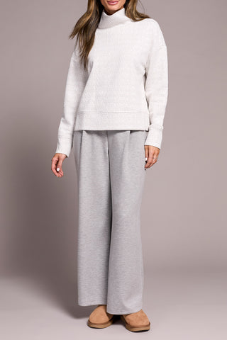 Person wearing a white and grey top with a funnel neck  with gray pants on a plain background.