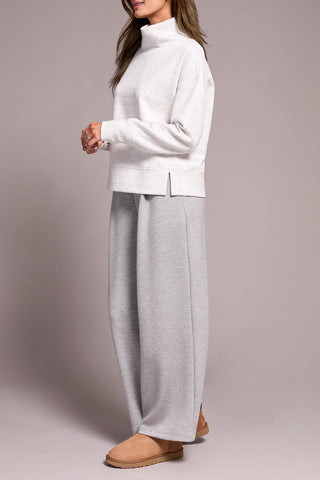 Person wearing a white and grey top with a funnel neck  with gray pants on a plain background.