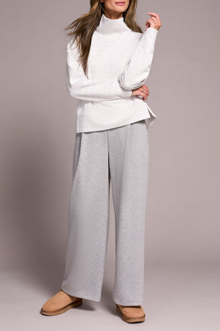 Person wearing a white and grey top with a funnel neck  with gray pants on a plain background.