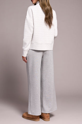 Person wearing a white and grey top with a funnel neck  with gray pants on a plain background.