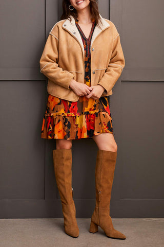 A woman wearing a beige long sleeve snap front reversible sherpa coat with a multicolored dress and brown boots.