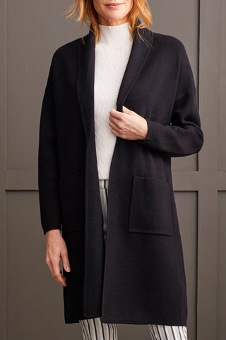 A woman wearing a black open-front coatigan with patch pockets over a white turtleneck sweater, paired with striped trousers.