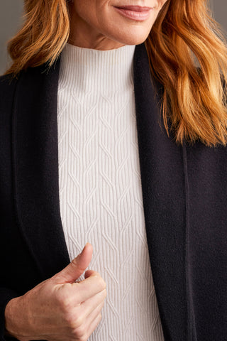 A woman wearing a black open-front coatigan with patch pockets over a white turtleneck sweater, paired with striped trousers.