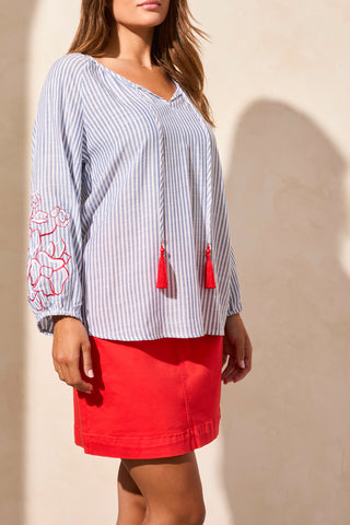 A woman wearing a blue and white striped blouse with red floral embroidery on the sleeves and red tassels, paired with a red skirt.