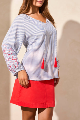 A woman wearing a blue and white striped blouse with red floral embroidery on the sleeves and red tassels, paired with a red skirt.