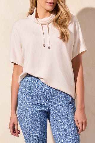 A woman wearing a heathered oatmeal funnel neck top with a drawcord at the neckline, paired with blue patterned pants.